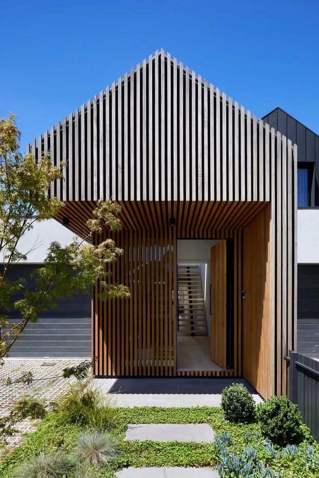 Front exterior view of a contemporary residential entry, featuring a strongly geometric gabled volume screened with closely spaced vertical timber battens. The timber screening continues across the recessed porch ceiling and walls, framing a glazed and timber front door. Dark horizontal garage doors, pale rendered walls, crisp black metal elements, and a landscaped path with low planting complete the facade. Bright, clear daylight emphasizes the rhythmic slatted design.