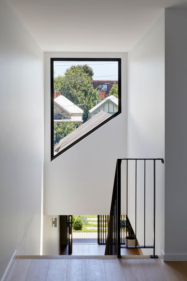 Bright minimalist stairway interior viewed from an upper landing. White walls and ceiling surround a large angular black-framed window, with a black metal balustrade enclosing the stair opening. Pale timber flooring continues across the landing, and a lower-level entry area with glazing and greenery is visible below. The geometric window frames views of neighbouring pitched-roof homes and mature trees.