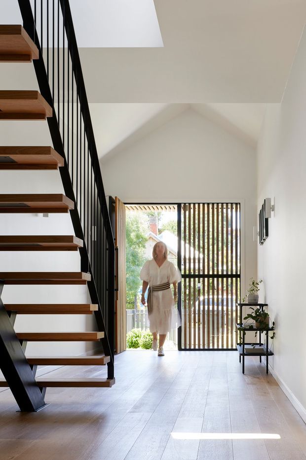 A bright, contemporary residential entryway with an open-riser timber staircase and slim black metal balustrade on the left. Pale timber flooring, white walls, and a high sloping ceiling create an airy interior. At the rear, a glazed entry door and vertical timber screening open toward greenery, while a small black console shelf with a potted plant sits along the right wall.