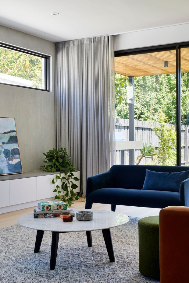 Bright contemporary living room with a deep navy sofa, round white marble coffee table, patterned rug, sculptural accent seating, and a minimalist white media cabinet. Floor-to-ceiling glazing opens toward a covered outdoor area, while full-height grey curtains and a trailing indoor plant add texture and softness.