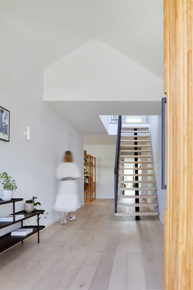 Bright minimalist residential hallway and stairway with pale timber flooring, white walls, an open-riser staircase with dark railing, and a large vertical timber-lined feature at the right edge. A slim console with plants and framed artwork adds light styling, while a person walks toward the stair area.