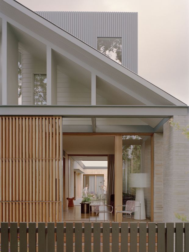 A contemporary multi-level residential facade viewed from outside, featuring a steeply sloping roof form, pale vertical metal cladding, white painted brickwork, horizontal weatherboard-like wall panels, and extensive glazing. A timber slat screen and dark vertical fence frame a covered entry courtyard, with glimpses of a refined interior lounge and an internal courtyard beyond.