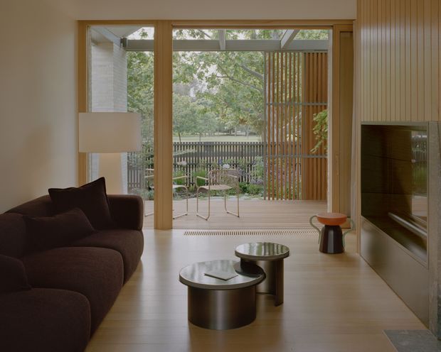 A warm, refined living room opening through full-height glazed doors to a small furnished outdoor terrace. The interior features pale timber flooring, a dark curved sectional sofa, minimalist cylindrical coffee tables, a floor lamp, and a large wall-mounted television within vertical timber-panelled joinery. The terrace includes a slatted timber screen, outdoor seating, and views toward mature trees and fencing.