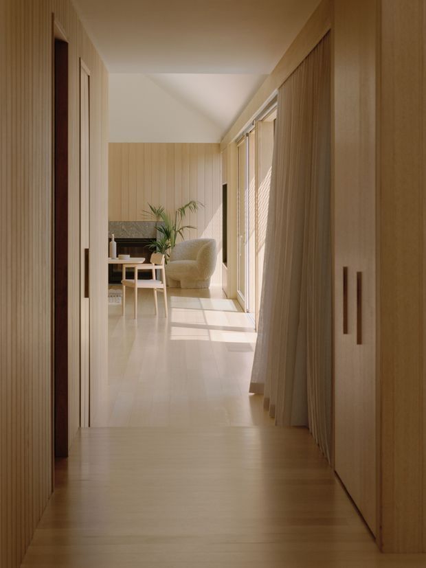 A warm, minimalist residential hallway lined with light timber wall panelling and built-in cabinetry. The corridor opens toward a softly furnished sitting area with a boucle armchair, small timber table, indoor plant, and dark stone or textured feature wall. Full-height glazing with sheer curtains brings in strong natural light, while the foreground has darker timber flooring transitioning to pale flooring beyond.