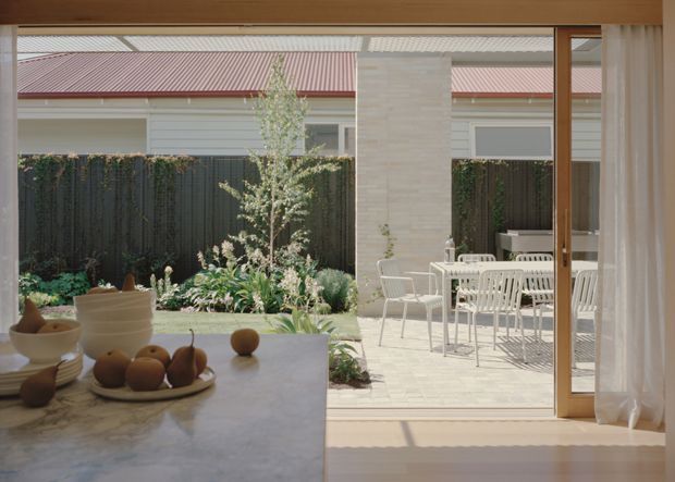 A bright contemporary interior opening onto a landscaped courtyard. The foreground shows a pale stone kitchen island or benchtop with bowls and fruit, while full-height glazed sliding doors lead to a paved outdoor dining area with white furniture. A pale stone-clad column, timber-toned window and door frames, and a neighbouring low-pitched corrugated roof are visible. The space has strong indoor-outdoor connection and abundant natural light.