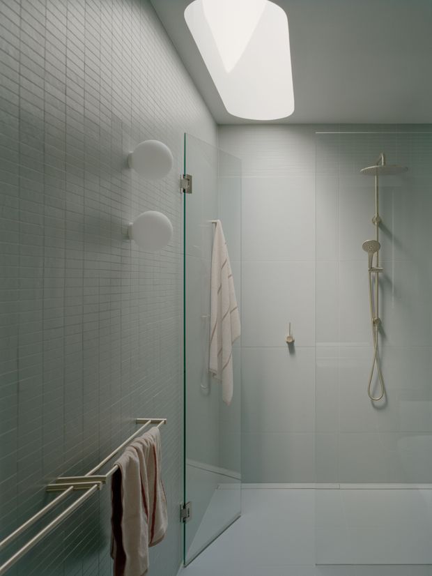 A compact contemporary bathroom with a walk-in glass shower enclosure. The room features pale blue-green small-format tiled walls, large light-toned wall panels, a white tiled floor, brushed metallic towel rails, and a streamlined shower system with overhead and handheld fixtures. A large sculptural recessed ceiling light provides soft illumination, creating a calm, clean atmosphere.