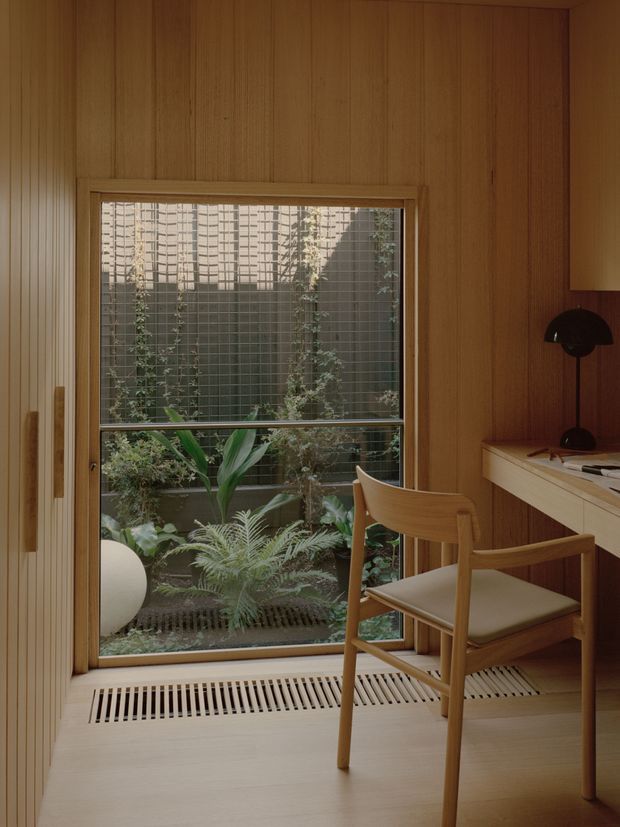 A warm, compact timber-lined home office or study nook beside a large window overlooking a planted courtyard. The space features vertical wood wall panelling, a built-in light timber desk with open shelving, a sculptural black desk lamp, and a simple timber chair with a light upholstered seat. A linear floor vent sits below the window, and lush indoor-outdoor planting is visible beyond the glazing.