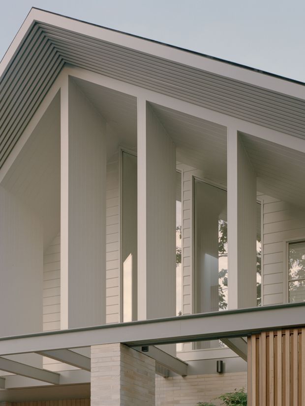 Close-up architectural exterior view of a contemporary residential facade. The composition features a deep upper-level covered porch or balcony framed by tall rectangular white columns, pale horizontal wall cladding, a strongly layered soffit with repeated linear detailing, and large glazed openings. A lower projecting canopy, pale brickwork, and vertical timber screening are visible below. The image emphasizes clean geometric forms, shallow overhangs, and restrained light neutral materials.