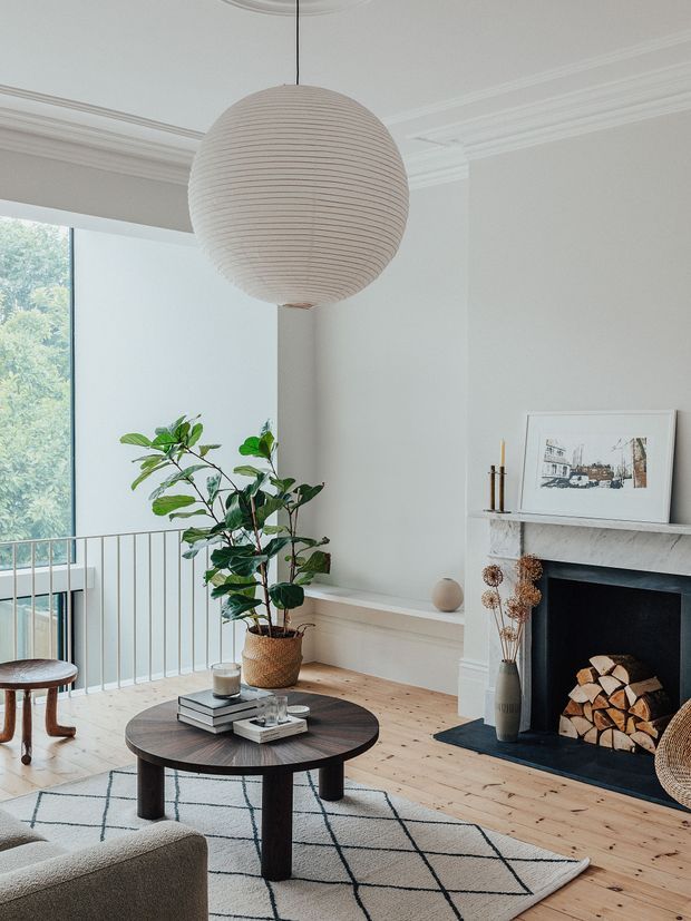 A bright, refined living room with high decorative moulded ceilings, white walls, pale timber flooring, and a large spherical pleated pendant light. The room features a dark round timber coffee table, neutral upholstered seating, a geometric cream-and-black rug, a tall indoor plant, and a traditional white mantel fireplace with stacked firewood. A large glazed opening overlooks leafy greenery and a slim metal balustrade.