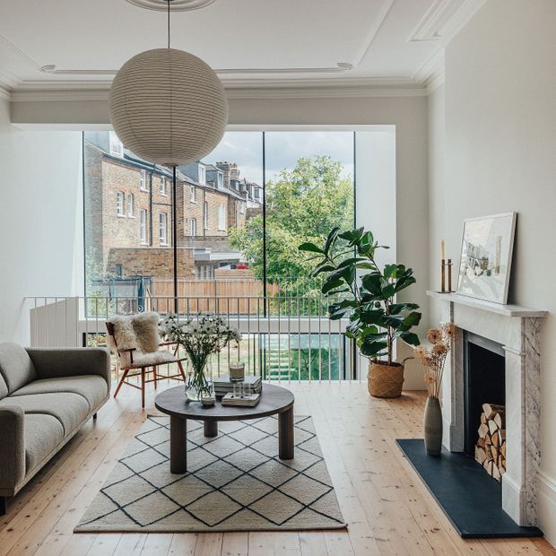 A bright, refined living room with a pale timber floor, large rear glazing and a slim balustrade overlooking neighbouring brick residences. The room features a neutral curved sofa, patterned rug, round dark coffee table, woven lounge chair, large indoor plant, a contemporary paper pendant, and a traditional white fireplace with a dark hearth and stacked firewood. Ornate ceiling cornices and recessed detailing add architectural character.