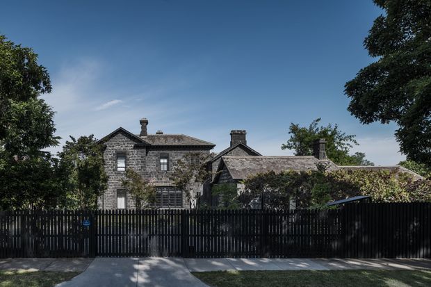 Exterior view of a substantial historic stone residence behind a tall black vertical-slat fence. The home has multiple gabled roof forms, dark grey slate-like roofing, masonry chimneys, and climbing vegetation, set among mature trees under a clear blue sky.