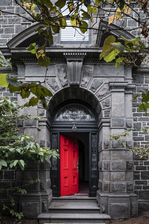 A close-up exterior view of an ornate historic entrance. The facade is built from dark grey masonry with heavy rusticated stone surrounds, classical cornices, pilasters, carved keystones, and arched detailing. A vivid red paneled timber door stands open beneath a decorative fanlight and sidelights, reached by a short flight of stone steps. Mature leafy vines partially frame the doorway.
