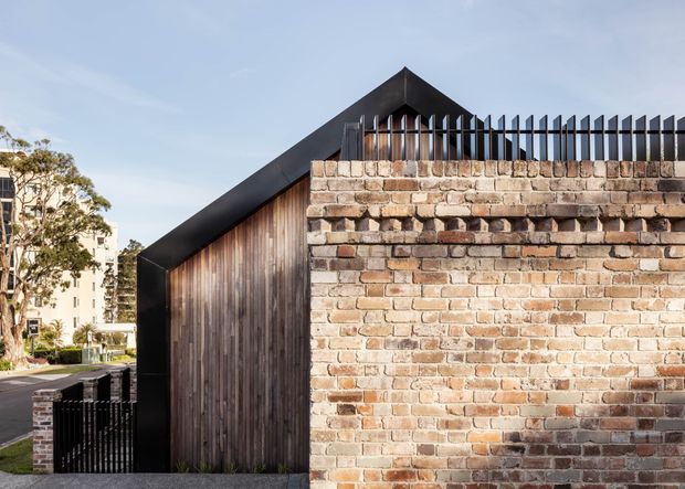 Exterior architectural view of a contemporary gable-roofed structure with dark metal roof edging, vertical timber cladding, and a prominent weathered brick boundary wall topped with a black metal picket fence. The image also shows a gated driveway or walkway and landscaped residential surroundings.