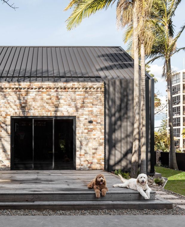 Exterior view of a compact contemporary building with a steeply pitched dark standing-seam metal roof, weathered multicolour brick facade, dark vertical metal cladding, and black-framed glazed doors. A timber deck and gravel-edged outdoor area occupy the foreground, with palm trees and neighbouring buildings visible around the structure.