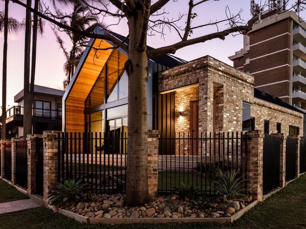 Twilight exterior view of a contemporary multi-level building with a dramatic steeply pitched glazed entrance volume, warm timber ceiling lining, exposed face-brick walls, and strong black metal framing. A brick gateway and low brick boundary wall sit behind a black metal fence, with established palms and a landscaped gravel garden in the foreground. Apartment balconies and rooftop services are visible in the background.