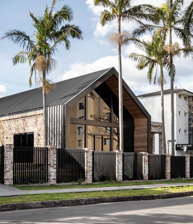 Street-facing view of a contemporary, single-storey gable-fronted building with a steep dark metal roof, exposed timber-lined gable, large glazed entry facade, and mixed brick and rendered masonry walls. A black vertical metal fence with brick piers fronts the property, with tall palm trees and neighbouring urban buildings visible around it.