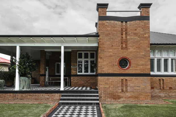 Front exterior view of a single-storey brick house with a prominent chimney-like brick feature, covered entry porch, black-and-white checkerboard paving, dark tiled roof, white columns, and louvered windows. The facade combines traditional brick detailing with crisp contemporary geometric elements.