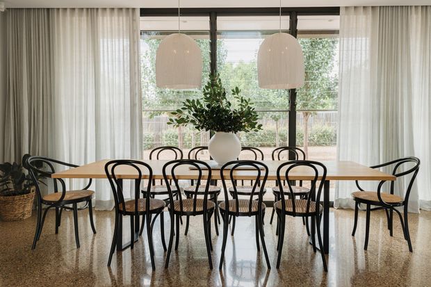 A refined dining room featuring a long light-wood dining table surrounded by black bentwood-style chairs with woven seats. Floor-to-ceiling sheer curtains frame large dark-framed windows, while two oversized pendant lights hang above the table. A white vase with leafy greenery forms the centrepiece, and the polished terrazzo-like floor reflects the furniture and light.