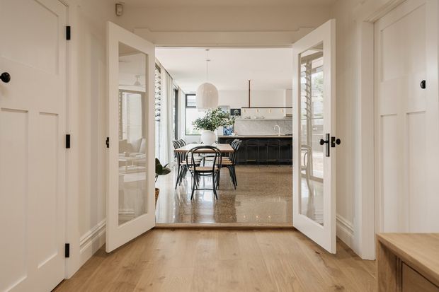View from a traditional hallway through open glazed double doors into a bright dining area and contemporary kitchen. The hallway has white paneled doors, decorative wall mouldings, and warm timber flooring. Beyond, a round dining table with black-framed chairs sits beneath a pendant light, with a dark kitchen island, pale cabinetry, stone-like splashback, and large windows bringing in natural light.