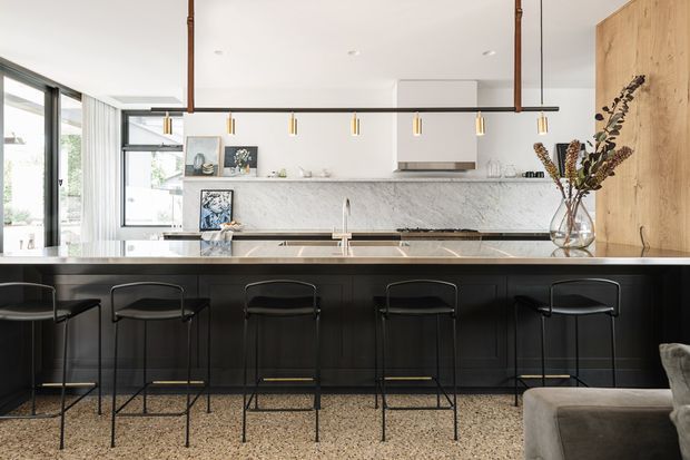 Bright, refined contemporary kitchen with a long black island and five slim bar stools, pale marble-look splashback, integrated stainless-steel appliances, warm timber cabinetry, and terrazzo flooring. Linear pendant lighting and expansive glazing create an airy open-plan setting, with part of a living area visible at right.