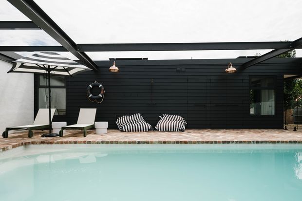 A contemporary outdoor pool area beside a dark horizontal-clad pavilion or pool house. The scene features a pale turquoise swimming pool, brick-paved pool surround, black steel pergola framing, outdoor lounge chairs, striped cushions, umbrellas, wall-mounted lighting, and a black-painted exterior wall with glazing.