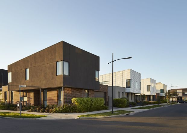 Street-facing view of a contemporary townhouse development, with a prominent two-storey corner dwelling clad in dark weathered metal and brick, alongside a row of similarly modern attached homes. The streetscape includes landscaped front setbacks, pedestrian paths, minimalist street lighting, and clear blue sky.