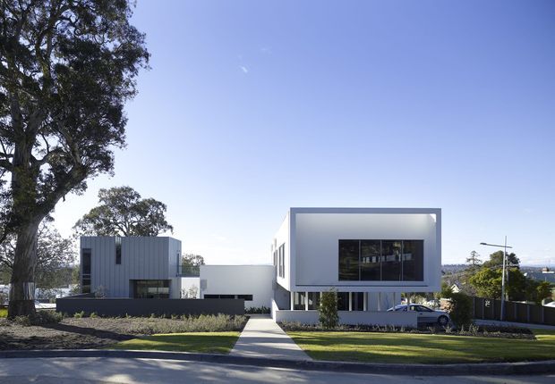 Street-facing view of a contemporary two-storey residence composed of crisp white and dark grey rectilinear volumes, with a projecting upper box, large dark-framed windows, a separate vertically ribbed grey side volume, landscaped front yard, and a central concrete pedestrian path. Mature trees, neighbouring homes, and a clear sky surround the property.
