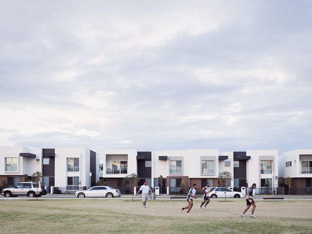 A row of contemporary multi-storey attached homes viewed across an open grass reserve. The residential facades use white and dark grey rectangular volumes, large windows, projecting entry canopies, balconies, and small front gardens. Several people and parked cars are visible in the foreground, beneath an overcast sky.
