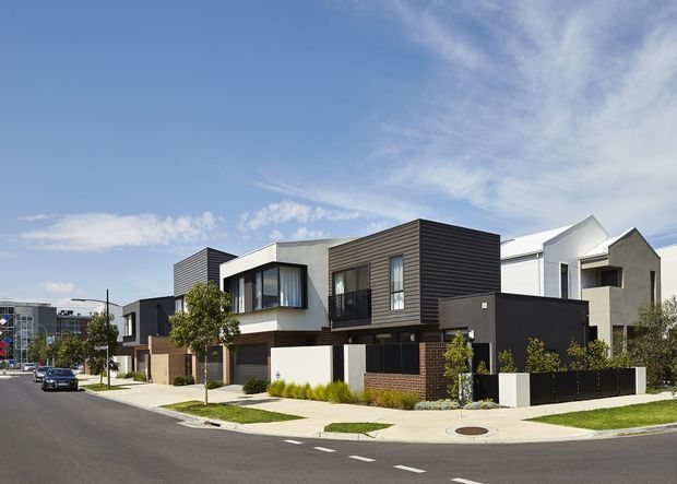 Street-level view of a contemporary suburban residential development featuring a row of attached and closely spaced multi-storey homes. The buildings have varied geometric volumes, flat and pitched roof elements, dark horizontal cladding, rendered white and grey walls, projecting upper-storey windows, garages, small landscaped front yards, and paved driveways. A broad asphalt street and clear blue sky frame the scene.