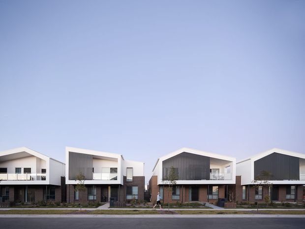 Street-facing view of a contemporary multi-unit residential development with repeated two-storey townhouse-like dwellings. The buildings feature steep gable roofs, white rendered frames, dark vertical cladding panels, exposed red-brown brickwork, upper-level balconies, and landscaped front setbacks along a paved street.