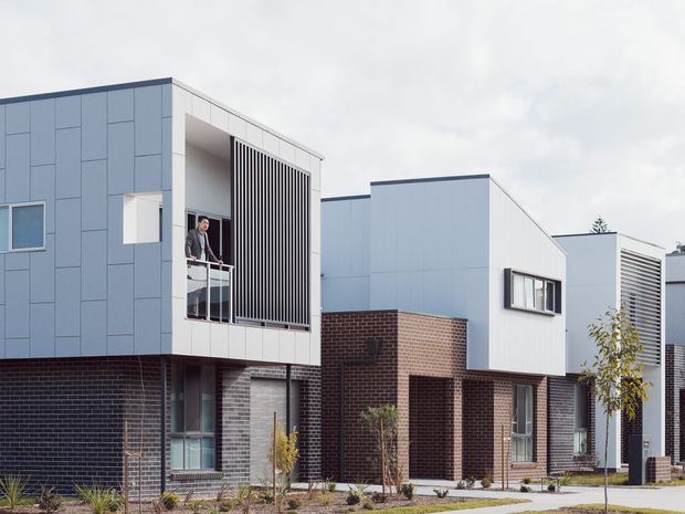 Exterior view of a contemporary multi-unit residential development with crisp white and pale blue-grey cladding, dark brick ground levels, projecting upper volumes, recessed balconies, and vertical privacy screens. Young landscaping and pedestrian paths line the frontage.