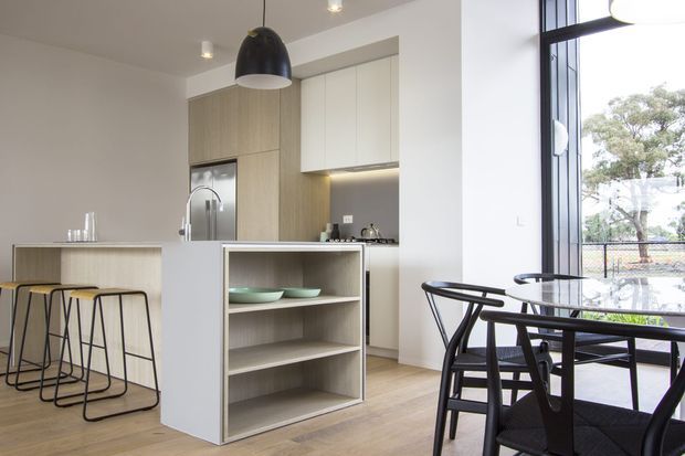 Bright contemporary open-plan kitchen and dining area with a compact island featuring open shelving and integrated seating. The kitchen has light timber-look cabinetry, white upper cabinets, a pale benchtop, stainless-steel fixtures, pendant lighting, and light timber flooring. A black dining table and chairs sit beside large black-framed windows overlooking greenery.