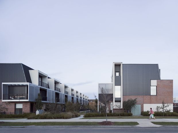 Street-level exterior view of a contemporary multi-unit residential development. The complex consists of attached townhouses or apartments with dark vertically ribbed metal-clad upper volumes, exposed red-brick lower walls, projecting balconies, and a narrow vertical window tower. Landscaped paths and planting separate the buildings from the street.