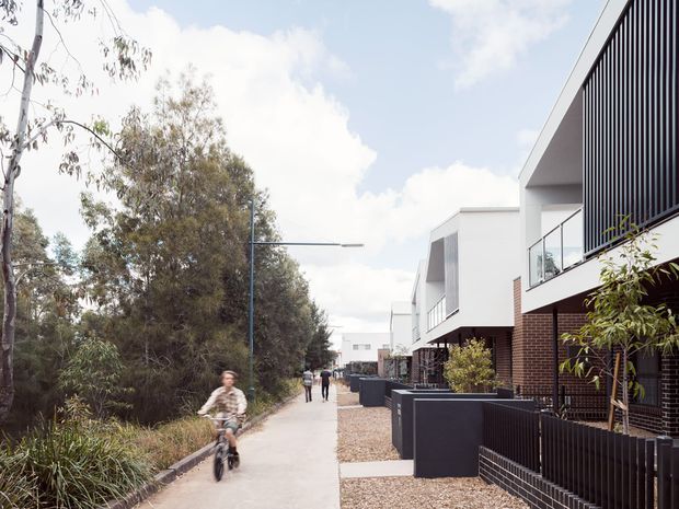 A contemporary residential streetscape with a paved pedestrian path running alongside a row of modern multi-level townhouses. The homes feature white rendered upper volumes, dark brick ground-floor facades, black vertical metal screening and balustrades, and small landscaped front setbacks. Native vegetation borders the opposite side of the path, with pedestrians and a cyclist visible in the distance.