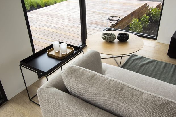 A bright, contemporary living room viewed from behind a light upholstered sofa. The room features pale timber flooring, a round light-wood coffee table, a black metal side table with tray and candles, a muted grey rug, and large black-framed corner glazing opening onto a timber deck and landscaped garden.