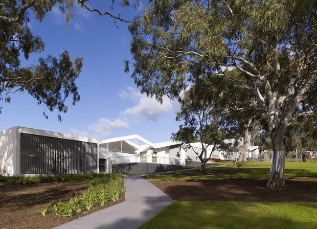 A low-rise modern building set within a landscaped suburban or institutional-style grounds. The white, rectilinear structure has flat roof forms, deep overhangs, open covered walkways, and a prominent dark vertical-louvre screen. A paved pedestrian path leads through lawn, garden beds, and mature eucalyptus trees toward the building.