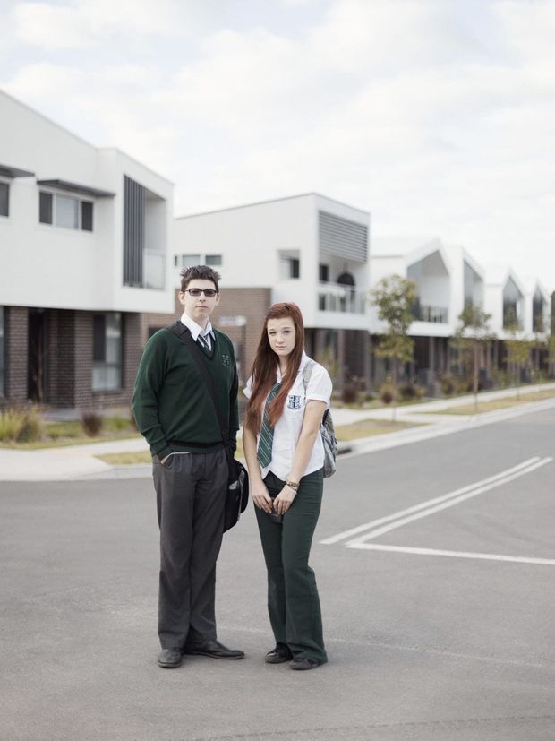 A street scene in a modern residential development, with two people standing in the foreground and contemporary white multi-unit homes lining the paved road behind them. The buildings feature boxy forms, flat roofs, dark brick lower levels, white rendered upper façades, vertical screening, balconies, and large rectangular windows.