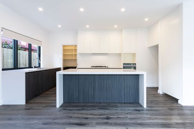 Bright contemporary kitchen with a large central island, white full-height cabinetry, dark vertically textured island cabinetry, dark timber-look flooring, integrated appliances, and a window-side benchtop with sink. Recessed ceiling downlights and minimal detailing create a clean, spacious interior.