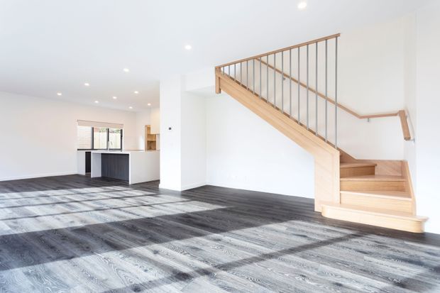 Bright open-plan residential interior featuring a timber staircase with slim vertical metal balusters, white walls and recessed ceiling lights. The staircase rises beside a large carpeted living area and leads toward a kitchen with a dark island visible in the background. Strong daylight enters through the windows, casting shadows across the grey patterned carpet.