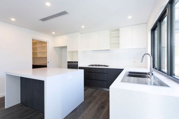 Bright contemporary kitchen with a large white island, dark vertically textured base cabinetry, white overhead cupboards, a white stone-look benchtop, integrated cooktop and oven, and a double-bowl sink beneath large black-framed windows. Dark timber-look flooring and a walk-in pantry or storage room are visible beyond the kitchen.