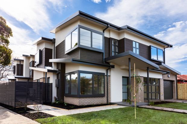 Exterior photograph of a contemporary multi-storey townhouse development with cream rendered walls, dark horizontal cladding, brickwork, extensive dark-framed glazing, projecting upper-level bays, flat rooflines, balconies or canopies, and landscaped front lawns with paved paths.
