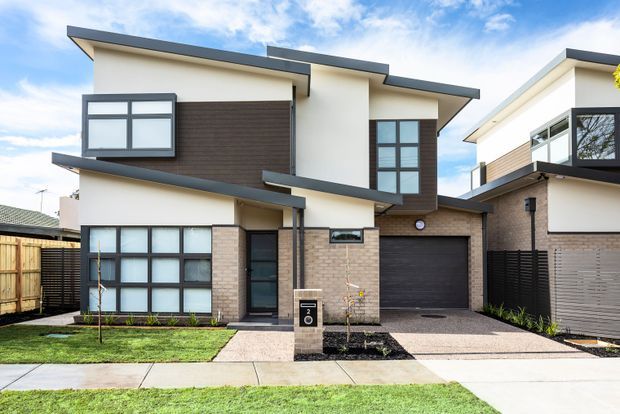 Front exterior view of a contemporary multi-storey townhouse with an angular low-pitched roofline, white rendered walls, dark horizontal cladding, light brickwork, large dark-framed windows, an attached single-car garage, front path, and small landscaped lawn.