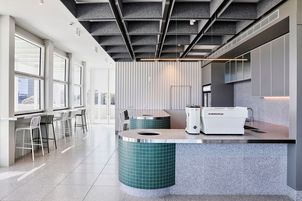 A bright contemporary commercial café or hospitality interior with a long coffee counter in the foreground. The counter has a rounded end clad in small teal-green tiles and a speckled grey benchtop and front. Behind it are coffee machines, dark grey fixed cabinetry, and open shelving. The space features large windows, pale tiled flooring, a vertically ribbed light wall, and an exposed dark acoustic ceiling grid with integrated services and linear lighting. Bar-height stools and tables line the window wall.