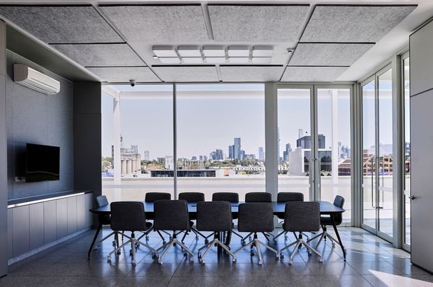 Bright contemporary commercial meeting room with a long conference table and rolling upholstered chairs, positioned beside full-height glazing overlooking an urban skyline. The room features a dark built-in media wall with wall-mounted screen and air-conditioning unit, grey acoustic ceiling panels, polished dark flooring, and a glazed corner with access to an outdoor terrace or balcony.