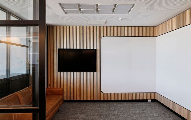 A compact contemporary commercial interior, likely a meeting or presentation room, with a large wall-mounted display set into a full-height vertical timber slat feature wall. The room has white curved-edged wall panels, dark carpet flooring, a brown leather built-in or sectional seat partly visible at left, extensive internal glazing with dark frames, and a perforated white acoustic ceiling with recessed square light panels.