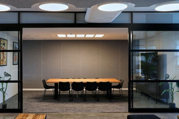 A contemporary commercial meeting room viewed through black-framed glazed partitions. The room contains a long timber meeting table with black chairs, grey carpet, textured grey wall panels, and recessed ceiling lighting. Indoor plants and adjacent office areas are visible through the glazing. The space has a clean, professional, moderately dramatic atmosphere with strong black framing and circular ceiling fixtures.