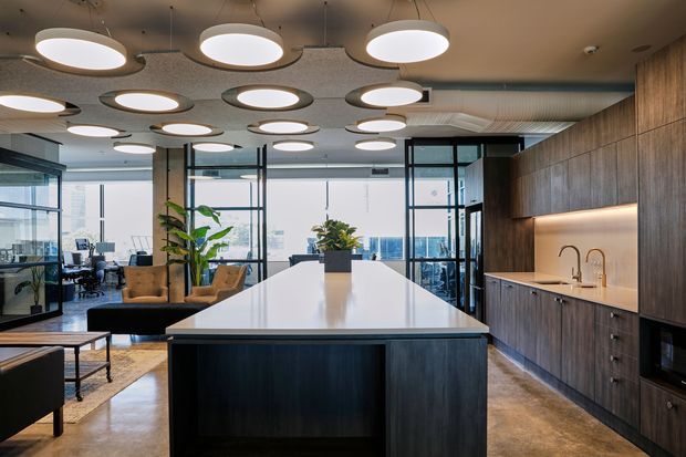 A contemporary commercial office breakout kitchen and lounge area with a large central island, dark timber kitchenette cabinetry, integrated appliances, and a pale benchtop. The space features polished concrete flooring, extensive black-framed glass partitions and windows, indoor plants, lounge seating, and numerous suspended circular pendant lights beneath an acoustic ceiling. A city view is visible through the glazing.