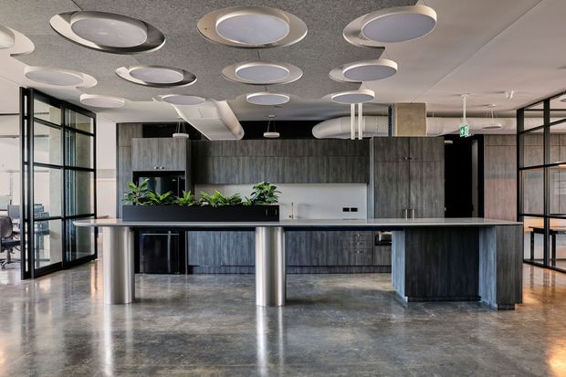 A modern commercial office breakout or kitchenette area with a long dark island counter, cylindrical metal supports, integrated cabinetry, and a planter strip. The space has polished concrete flooring, dark timber-look joinery, extensive black-framed glazing, and a distinctive ceiling fitted with numerous circular acoustic light fixtures. Open workspaces and glass partitions are visible around the central fit-out.
