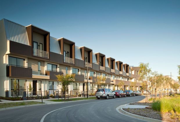 Exterior street view of a contemporary multi-storey townhouse development, featuring repeated projecting upper-level volumes, dark metal balcony balustrades, light rendered walls, vertical metal screening, and ground-level glazing. The homes front a curved paved street with landscaped verges and young trees.