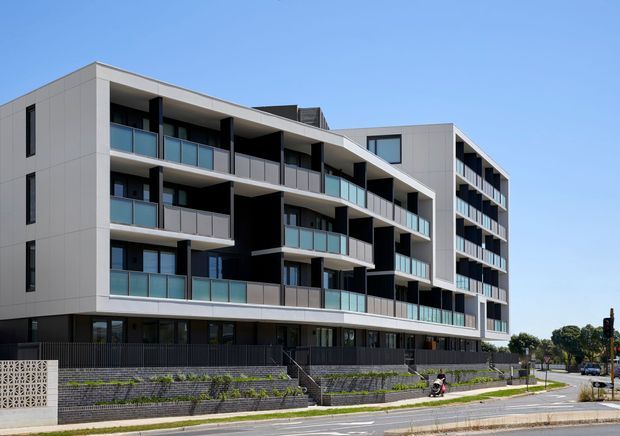 Exterior view of a contemporary multi-storey apartment building with projecting balconies, pale panelled facade, dark recessed sections, and blue-tinted glass balustrades. The building sits above landscaped retaining walls along an urban street.